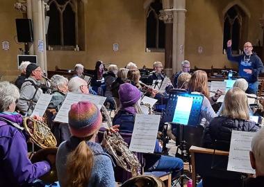 music creator Peter Falconer is standing and conducting the Band of the Surrey Yeomanry, who are seated and playing their instruments