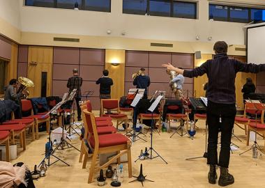 composer leading a workshop by having the brass band players face the walls while playing their instruments