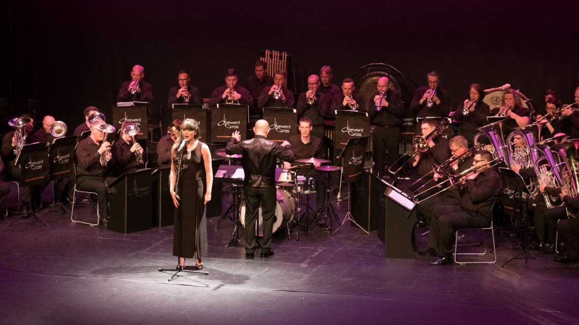 A woman at the centre of the stage singing, with a band behind playing various brass instruments. All are dressed in black and under a purple light. 