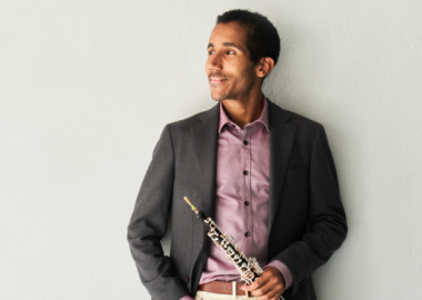 smiling man wearing pink shirt and grey jacket, holding oboe