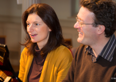 Smiling woman wearing yellow cardigan seated next to laughing man wearing glasses, both sat at piano